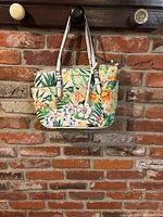 Front view of the tropical floral Dana Buchman purse hanging against a brick wall, showing the floral design, white straps, and front pocket with flower charm.