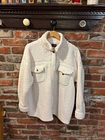 White fleece jacket with button-up front and two buttoned chest pockets hung on a black hook against a brick wall background, showing front view.