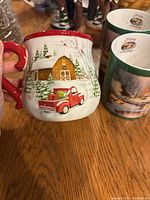 Side view of a holiday mug showing a red truck carrying a Christmas tree near a barn and snowy pine trees.