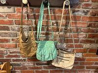 Three satchel bags hanging on hooks against a brick wall; colors are teal, sandy brown, and beige