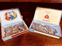 Two open cigar boxes filled with assorted coins, one Montecristo brand, the other a decorative Cuban box