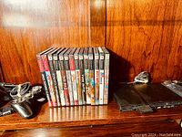 PS2 Slim console, two controllers, and 13 game cases lined up upright on wood shelf.