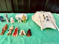 Top-down photo of all figures and vehicles laid on green surface: white Rebel Armoured Snowspeeder vehicle, white Escape Pod vehicle, 12 Star Wars action figures from 1977-1980 Kenner line including droids, heroes, and creatures.