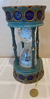 Front view of ornate hourglass music box with turquoise and gold coloring, zodiac symbols, and white sand inside the glass.