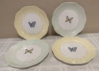 Four Lenox dessert plates arranged side by side, showing one blue butterfly on yellow rim plate and one yellow and black butterfly on green rim plate.