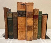 Eight antique and collectible books lined up vertically, showing varied aged and leather bindings with titles visible on spines.