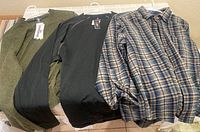Three men's shirts on hangers laid out on table: Pendleton plaid button-up, black L.L. Bean Polartec pullover, olive green Smartwool sweater