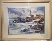 Framed watercolor print showing a coastal scene with lighthouse, ocean, rocks, and buildings in a beige wood frame.