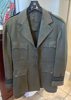 Front view of the olive green military coat jacket hanging on a hanger, showing buttoned front with four pockets and eagle buttons.