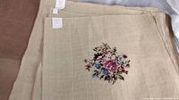 Stack of beige needlepoint canvases with central bouquet motif and corner labels