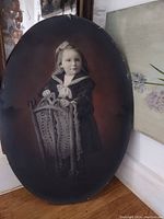 Oval black and white photograph of a young child standing behind an ornate carved chair, circa 1900.
