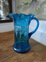 Front view showing the bright blue glass, ruffled rim, handle, and handpainted floral decoration with some wear.