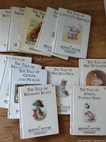 Twelve hardcover Beatrix Potter books with white dust jackets, laid flat on wooden surface. Titles include 'The Tale of Benjamin Bunny', 'The Tale of Two Bad Mice', 'The Tale of Jemima Puddle-Duck' and others. Visible wear on jackets from age and use.