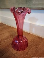 Cranberry glass vase shown standing on wood floor, approx. 8 inches tall, bulbous ribbed base with tall flared ruffled neck, translucent cranberry glass color.