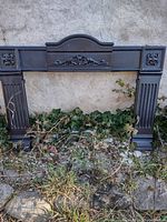 Front view of the black heavy iron fireplace mantel showing decorative floral scrollwork and expandable side legs with fluted detailing.