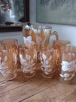 Full set includes one pitcher and six glasses with white floral decorations on marigold carnival glass.