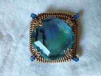 Front view of a large square brooch with a central multi-faceted blue rhinestone and gold-tone coiled wire frame with blue accent rhinestones at corners.
