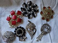 Top view of seven vintage brooches laid on white fabric, showing front details including colored stones and rhinestones.