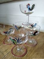 Photo of six small cocktail glasses grouped together showing handpainted multicolor rooster designs and pink rings on base