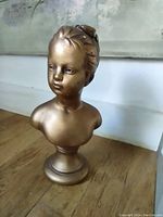 Photo showing gold-painted plaster bust of a young girl with some paint loss on top, about 8 inches tall, on wooden surface against a wall.