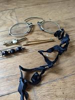 Vintage eyeglass frames, three vintage pins, and a black silk ribbon accessory arranged on wooden floor.
