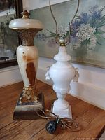 Two antique heavy lamp bases made from marble or onyx shown side-by-side on a table against a backdrop with floral paintings. Taller lamp approx 18 inches, smaller lamp shows some chips.