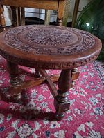 Full view of the round wooden stool showing the carved leather top and three turned wooden legs on a patterned rug.