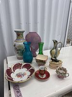 Lot with ten porcelain and glassware pieces shown on a white surface outside against a corrugated metal background, showing vases, pitcher, plate, cups and trinket holder.