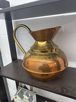 Photo showing the front and handle side of the large copper and bronze pitcher on a dark shelf against white wall background.