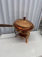 Side angle view of copper chafing dish showing wooden handle and decorative stand with fuel holder beneath.