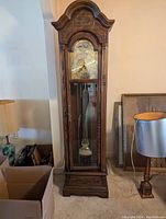 Front view of the full vintage grandfather clock showing the brass face, carved wood case and glass door allowing view of pendulum and weights.