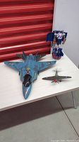 View of all three items on table: large blue camo fighter jet, small vintage WWII plane, and a standing red, blue, and silver transforming robot figure.