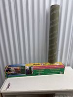 Photo of boxed Roll-O-Puzz puzzle roll-up mat standing vertically beside the box on a white surface against a corrugated metal backdrop.