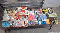 Photo showing sewing magazines, packaged sewing machine needles, and zigzag stitch accessories metal tin along with several packaged sewing hardware items.