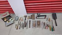 Wide view of assorted vintage hand tools laid out on concrete including chisels, trowels, scrapers, pliers, screwdrivers, metal box, and lamp cage.