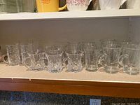 Row of 21 clear glass cafe coffee mugs with handles and cut designs, displayed on shelf.