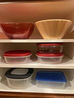 Shelf with assorted kitchen containers and bowls: wooden salad bowl, red plastic mixing bowl, and multiple clear plastic containers with colored lids.