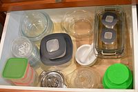 Drawer filled with various clear glass Pyrex mixing bowls and rectangular serving dishes with lids, plastic containers with multiple lids in gray and green, small glass bowls, a metal strainer, and white ceramic spice grinder bowl with pestle.