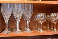 Photo showing two sets of crystal wine glasses on wooden shelf: six smaller clear glasses without pattern, six larger crystal glasses with diamond cut pattern.