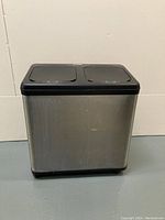 Front view of the rectangular stainless steel dual compartment trash can with black base and slightly worn surface.