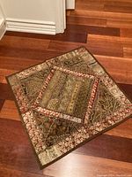 Square handmade cushion cover with ornate patchwork in green, brown, and beige tones with decorative border, placed on wood floor.