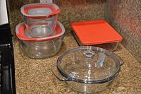Photo showing two nested square clear Pyrex glass baking dishes with red rubber lids and one rectangular dish with flat red plastic lid and one round dish with glass lid on countertop.