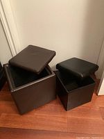 Two dark brown faux leather storage ottoman cubes, lids removed showing internal storage space, placed on wood floor against white wall.
