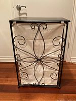 Front view of laundry hamper, showing black iron wire frame with intricate scroll designs and beige canvas liner.
