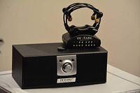 Photo showing one TV Ears wireless headset earpiece resting on top of the transmitter base unit against a neutral background.