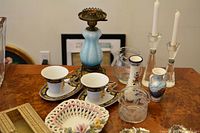 Overview of lot showing porcelain cookie bowl with floral detail, small fishbowl, gold napkin holder, two ceramic vases, two crystal candle holders, two teacups with saucers, and antique blue crystal lamp with bronze base (missing shade).