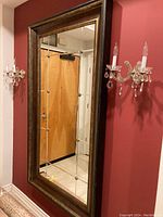 Front view of the large vintage floor mirror, showing full rectangular shape with dark burgundy and antique gold wood frame, mounted on red wall between two sconces.