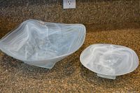 Photo showing two white decorative glass serving bowls on a kitchen countertop, clearly displaying their size difference, squarish shape, and marbled white and gray pattern with scissor-cut edges.