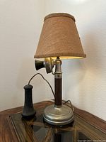 Front angle view of the candlestick telephone lamp showing the black handset corded to the metal base and the beige fabric lampshade.