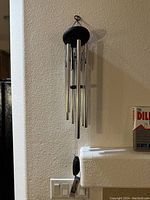 Full view of the wind chime hanging against a wall, showing silver metal tubes, black wooden top and wind catcher.